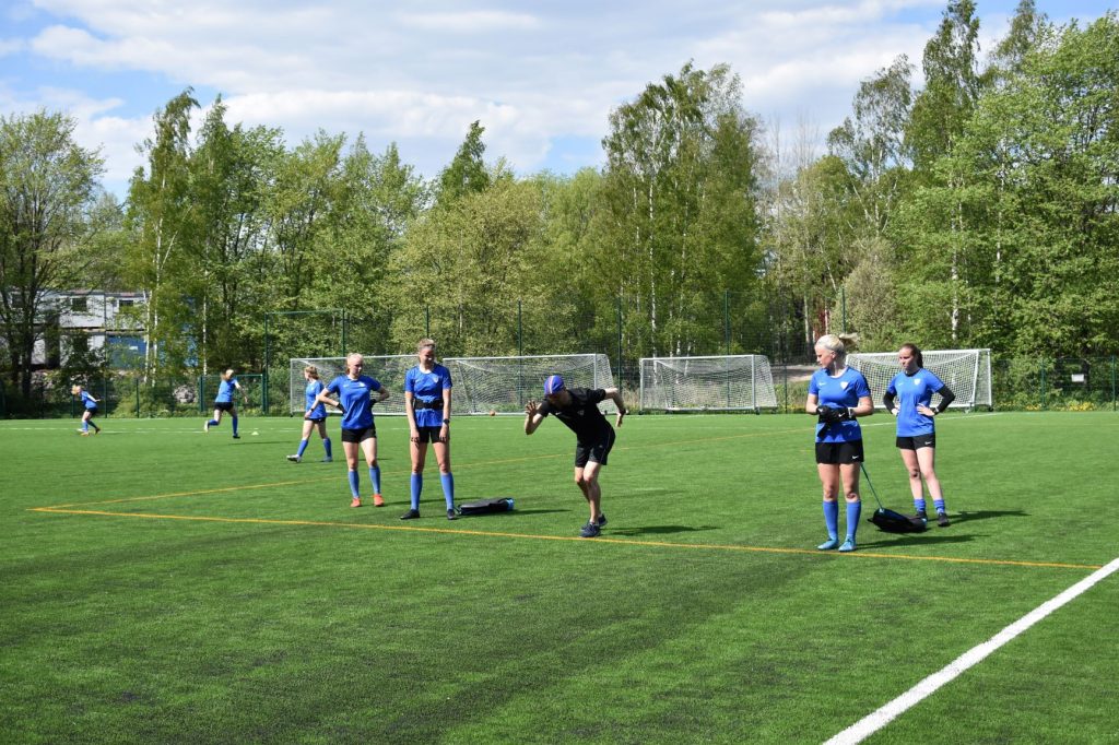 Kesäisellä jalkapallokentällä jalkapalloilijat katsovat, kun valmentaja antaa ohjetta juoksuasennossa. Taustalla pari pelaajaa potkii palloa.
