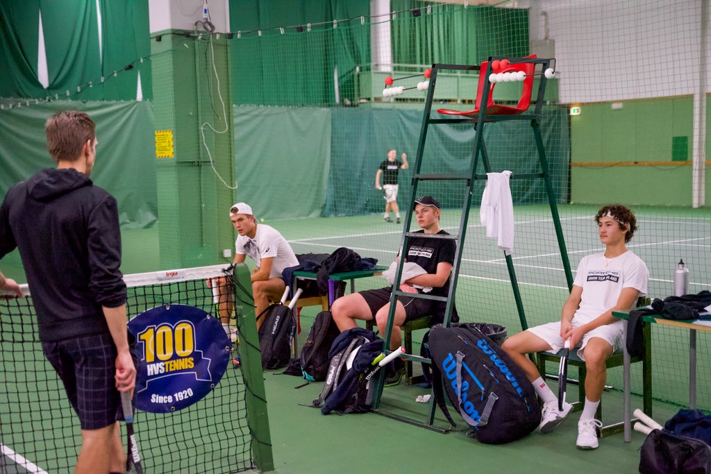 Tenniskentällä kolme pelaajaa istuu kentän laidalla tuoleilla ja katsoo kohti valmentajaa, joka seisoo verkon vieressä