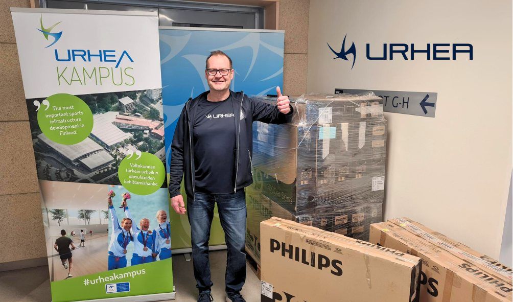 Mika Saari nojaa televisiopinoon ja näyttää peukaloa ylöspäin, taustalla Urhea-logot